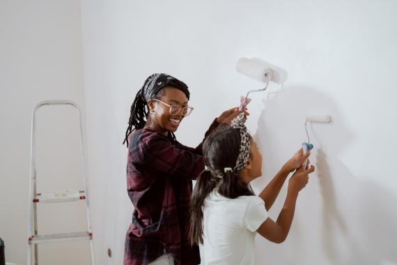 Busy mom spends time with cheerful daughter, engages her to help, together they painting the walls of the apartment under renovation, apply white paint with rollers 